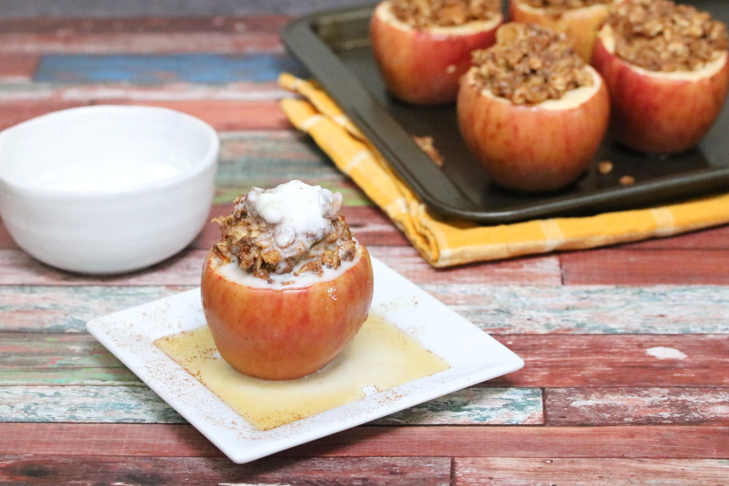 Apple stuffed with oatmeal and topped with whole 30 whipped cream on a white plate with maple syrup on it and more apples in a pan behind them.