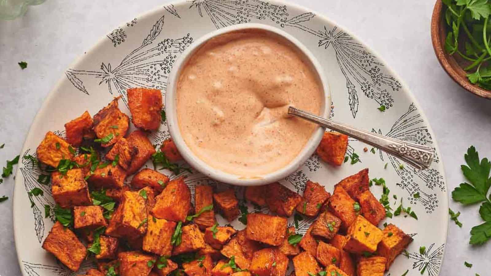A plate of roasted sweet potato cubes garnished with herbs, served with a bowl of creamy dipping sauce and a spoon.