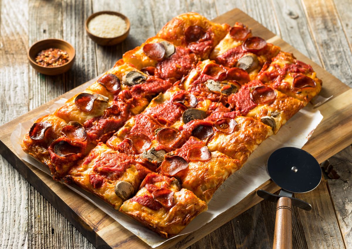 Rectangular Detroit-style pizza with pepperoni, mushrooms, and tomato sauce on a wooden board, with a pizza cutter and bowls of grated cheese and crushed red pepper nearby.