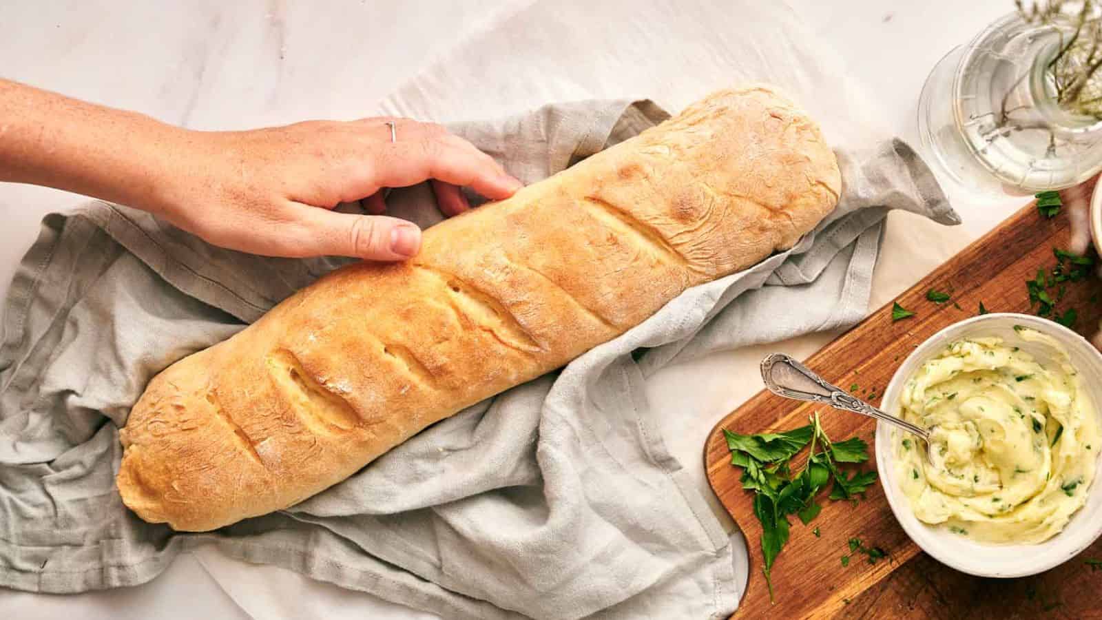 A hand reaches for a loaf of baguette on a cloth, next to a cutting board with a bowl of herb butter and fresh parsley.