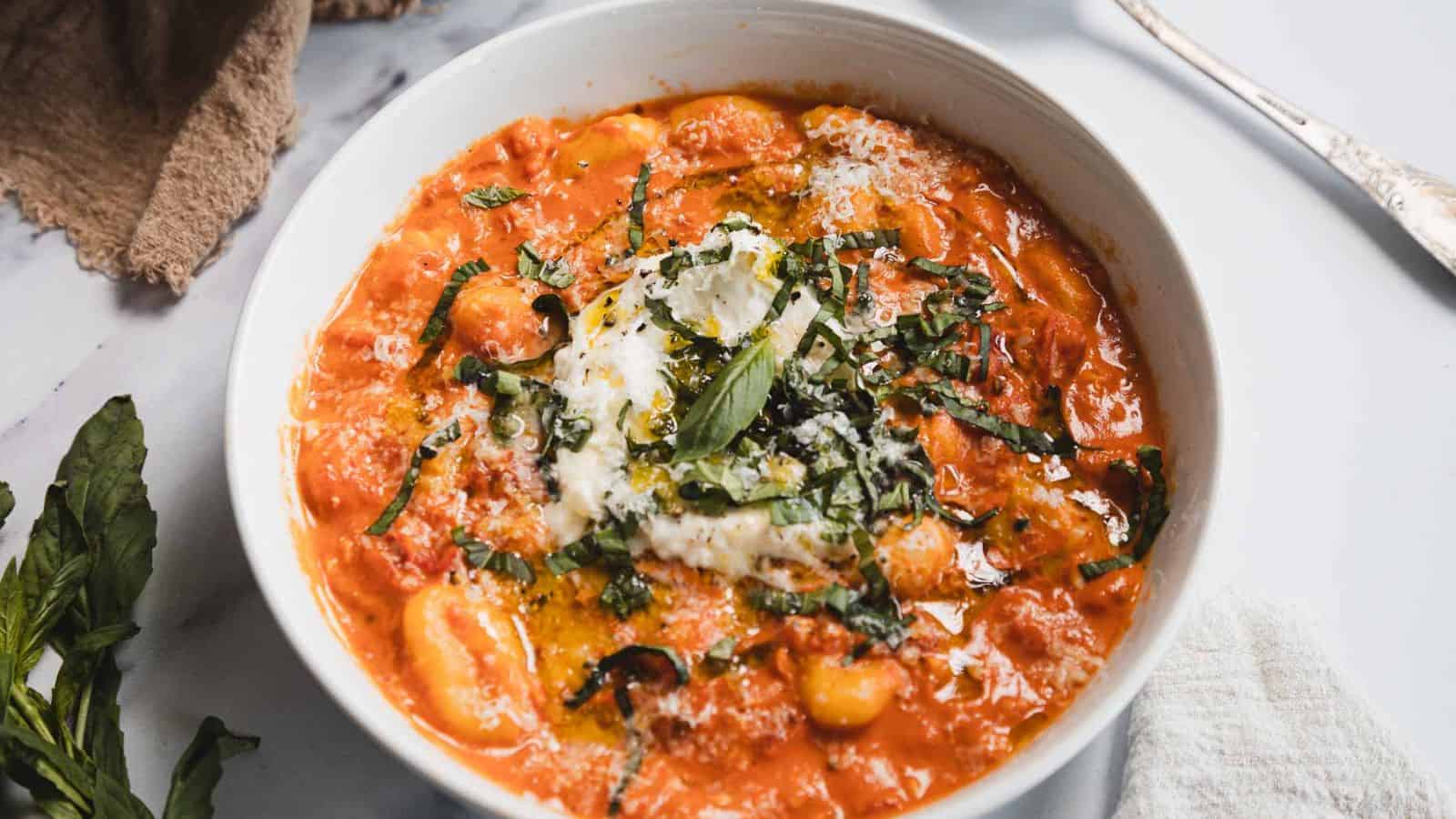 A white bowl filled with gnocchi in a creamy tomato sauce, topped with melted cheese, chopped basil, and a drizzle of olive oil.