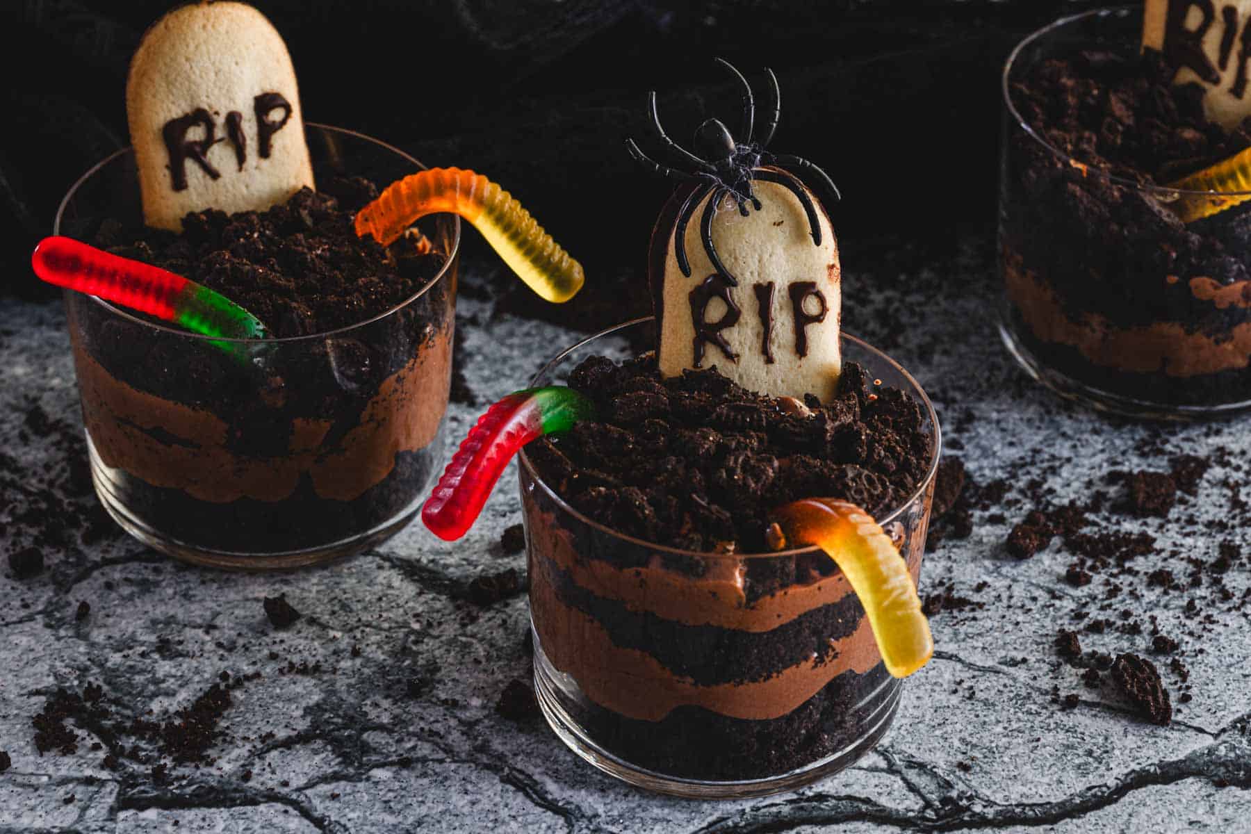 Three glass cups with layered chocolate pudding and crushed cookies, topped with gummy worms and a cookie marked RIP to resemble a graveyard, with a plastic spider decoration.