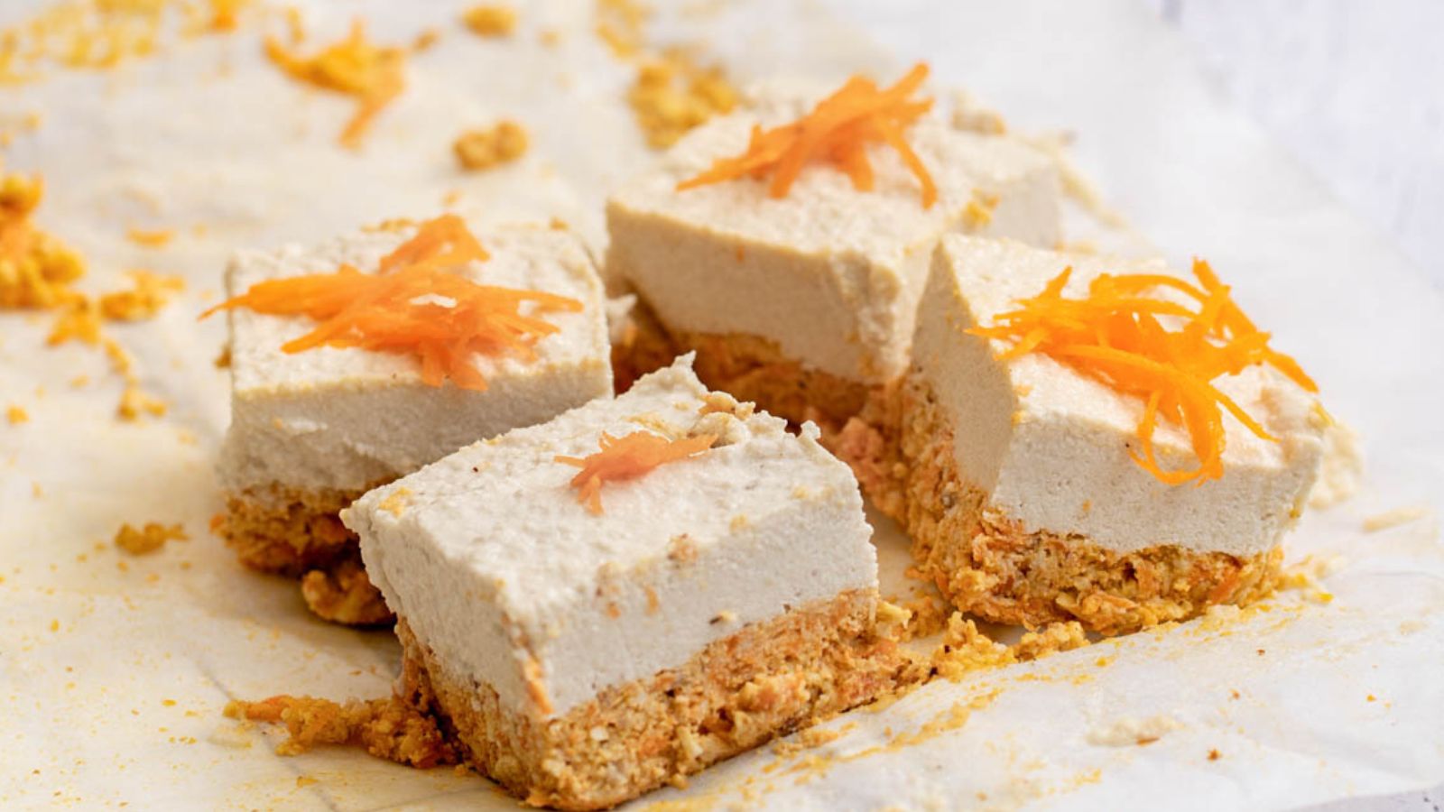 Four square pieces of cheesecake with a crumbly base and creamy topping, each garnished with shredded carrot, are arranged on a sheet of parchment paper.