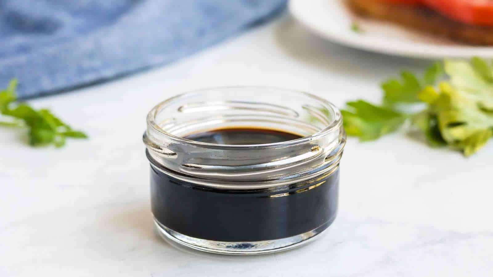 A small glass jar filled with dark liquid, likely balsamic vinegar, is placed on a light surface with some herbs and a plate blurred in the background.