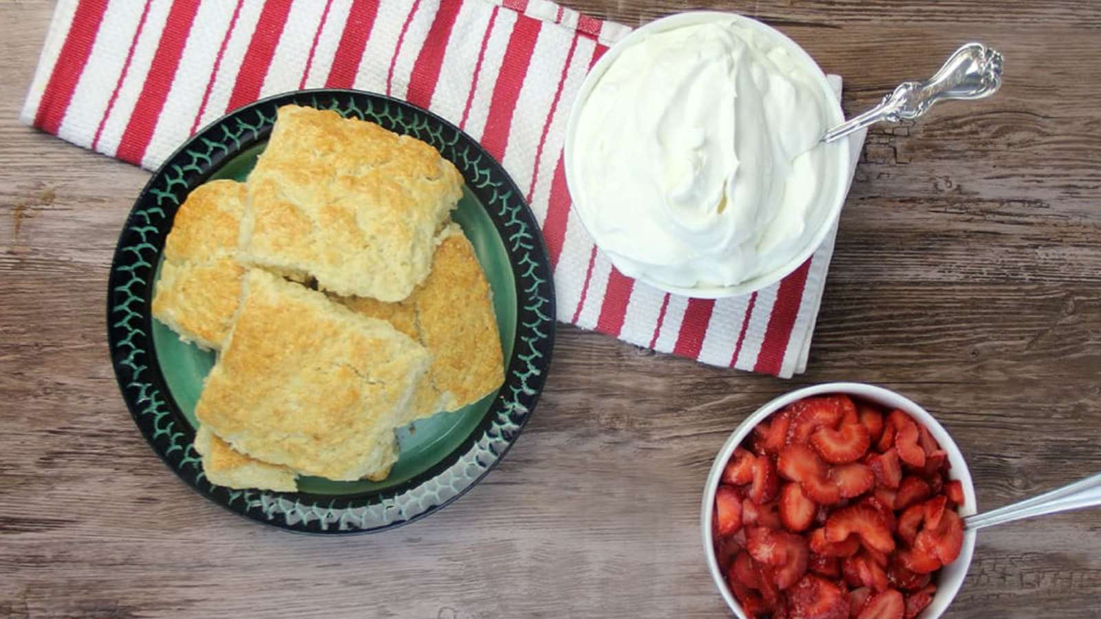 A plate of biscuits, a bowl of whipped cream with a spoon, and a bowl of sliced strawberries are arranged on a wooden table with a red and white striped cloth.