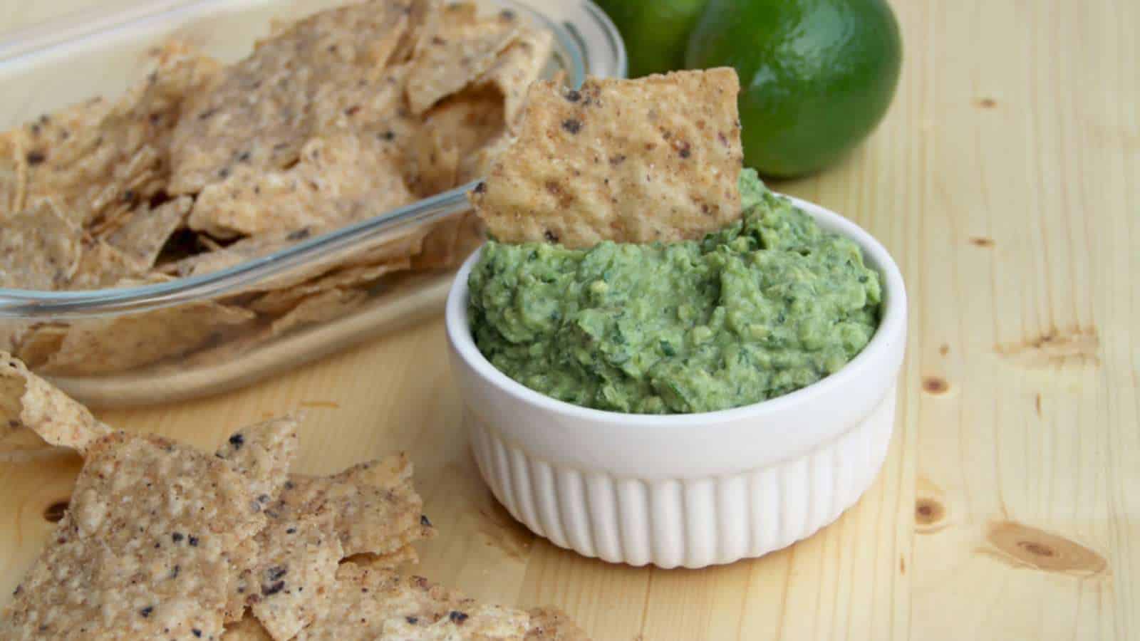 A bowl of guacamole with a tortilla chip dipped in it, surrounded by more chips and a lime on a wooden surface.