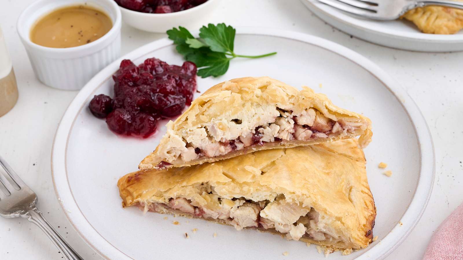 A plate with a sliced puff pastry turnover filled with turkey and cranberry sauce, served with a side of cranberry sauce and parsley garnish.