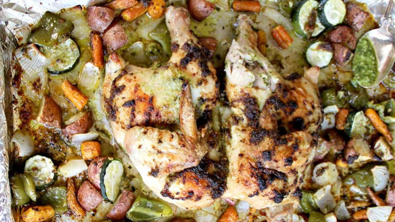 Roasted spatchcock chicken on a foil-lined tray surrounded by cooked carrots, zucchini, onions, potatoes, and green bell peppers.