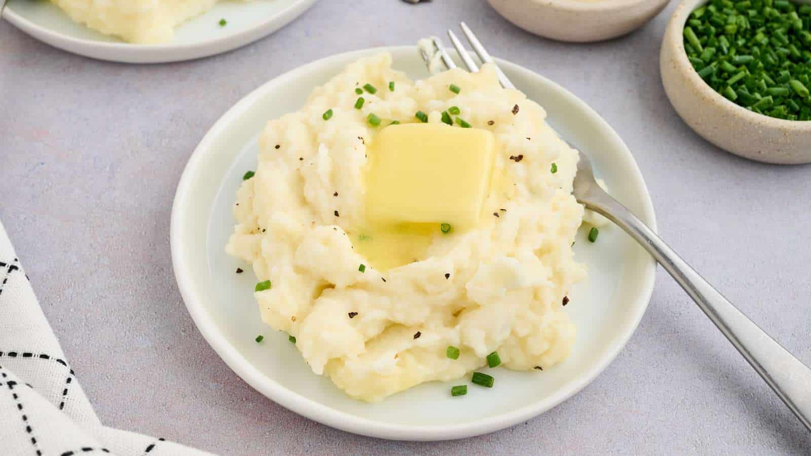 A plate of mashed potatoes topped with a pat of melting butter and chopped chives, with a fork on the side.