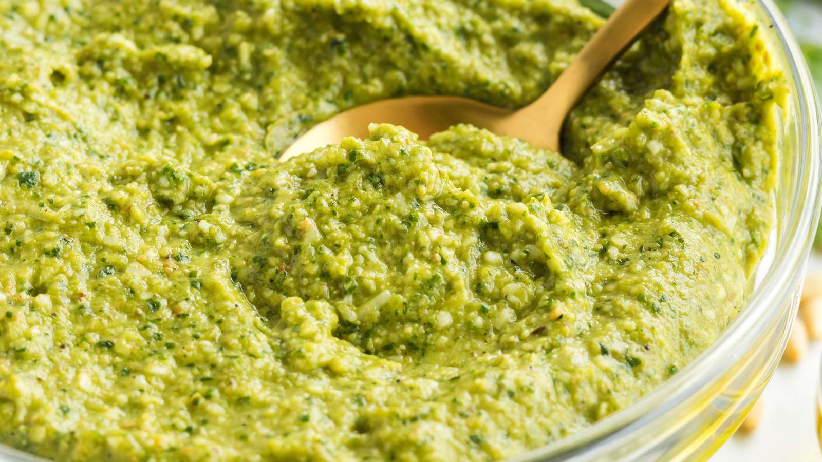 A close-up of a bowl filled with green pesto sauce, with a gold spoon partially submerged in the mixture.