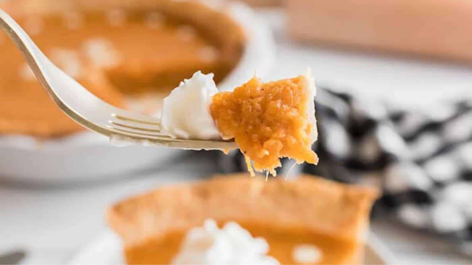 A fork holds a bite of pumpkin pie topped with whipped cream, with a pie and a slice visible in the blurred background.