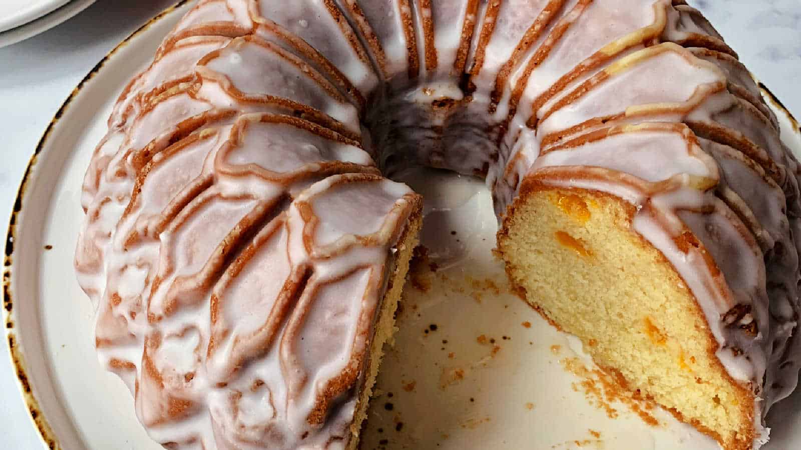 A glazed bundt cake with a slice removed, showing the moist interior and pieces of fruit inside, on a round plate.