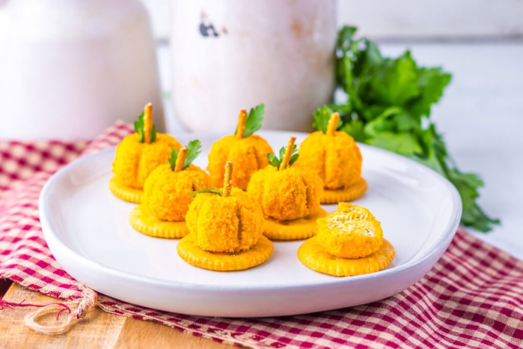 Round crackers topped with small, orange cheese balls shaped like pumpkins, garnished with parsley and pretzel stick stems, arranged on a white plate.
