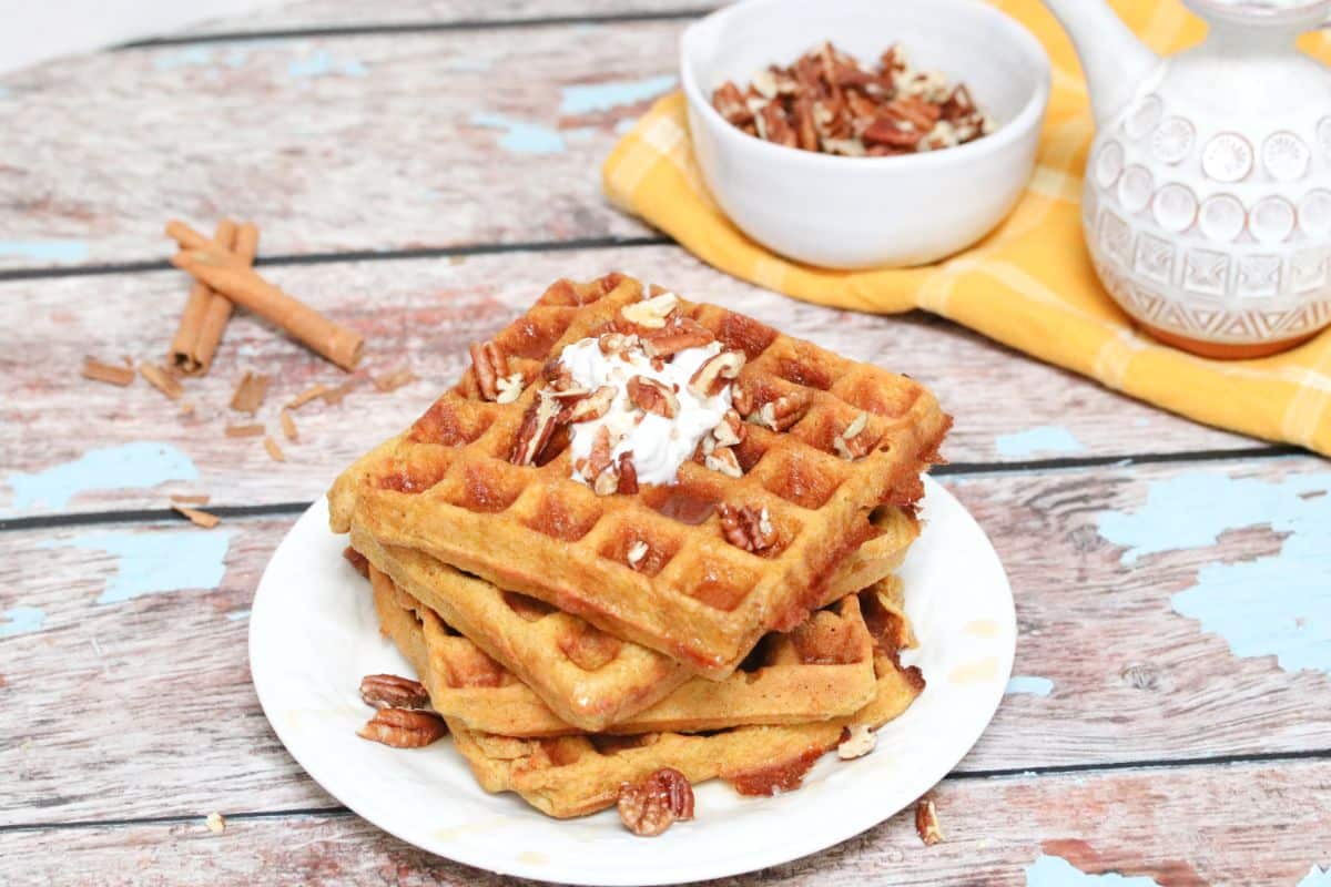 three gluten-free pumpkin waffles stacked on a white plate with coconut cream, maple syrup and pecans on top. A waffle maker beside the plate and a bowl of pecans and syrup behind it