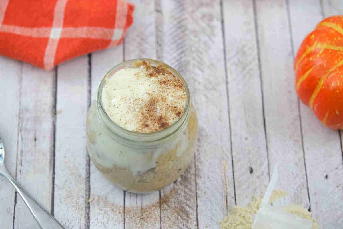 Mason jar with pumpkin spice overnight oats with cream cheese frosting on top and an orange linen and decorative pumpkins behind it.