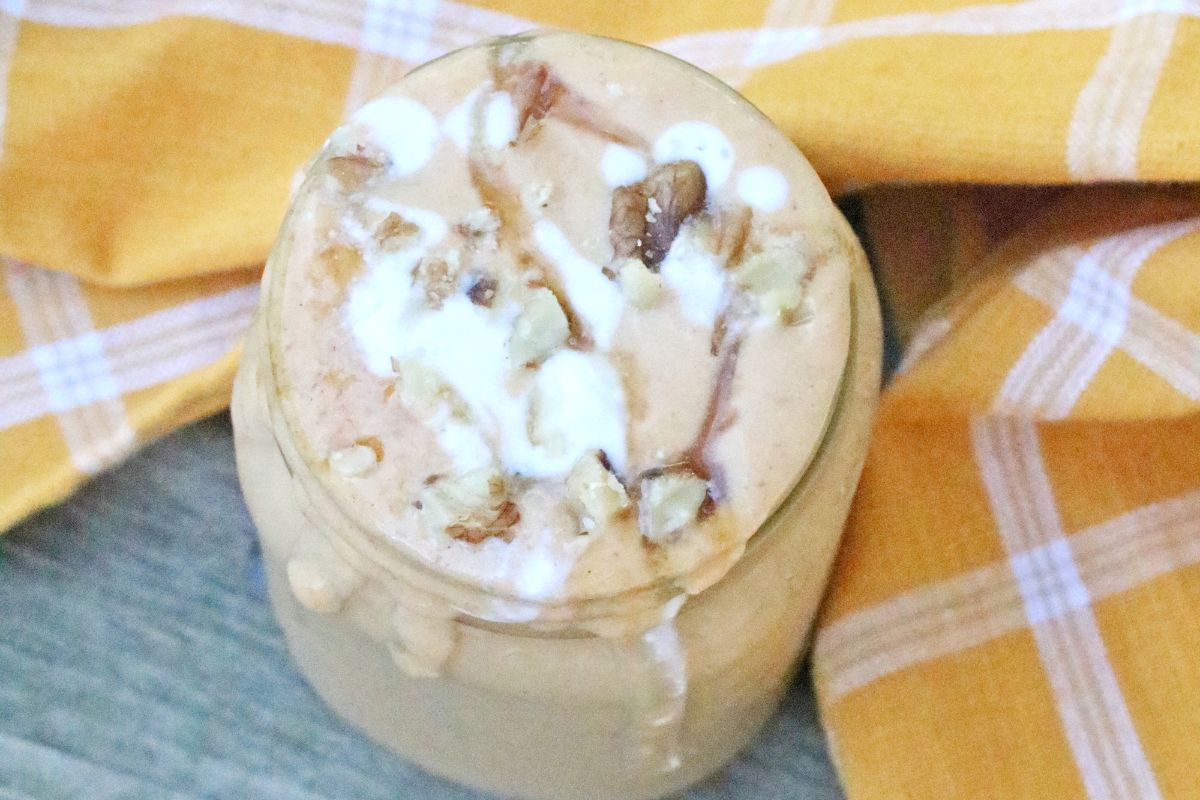 Mason jar with light orange pumpkin smoothie tipped with coconut whip cream, nuts and a blue straw spilling down the side.