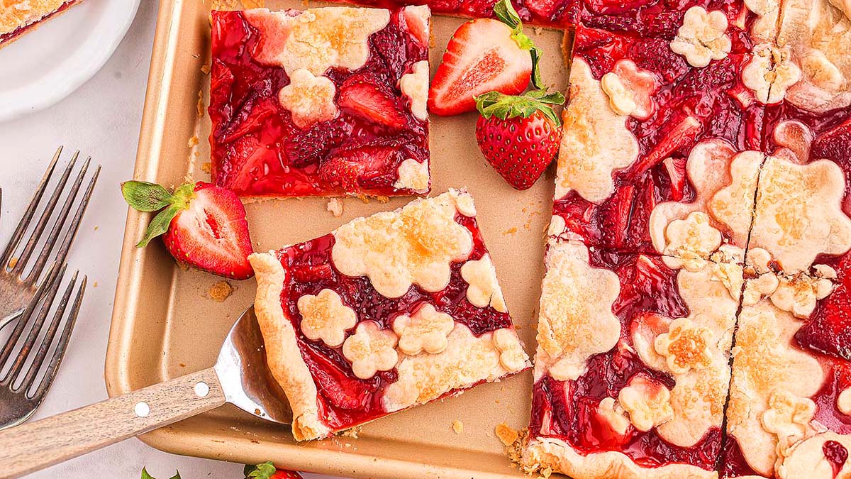 A rectangular strawberry pie with a decorative flower-shaped crust is cut into squares on a baking sheet, surrounded by fresh strawberries and forks.