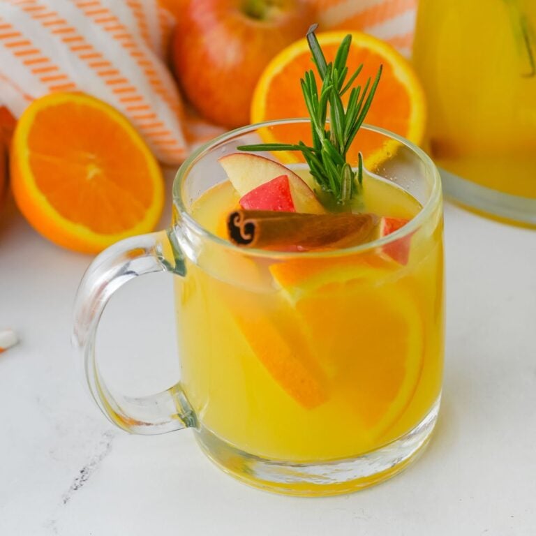 A clear glass mug filled with yellow citrus drink, garnished with orange slices, apple pieces, a cinnamon stick, and a sprig of rosemary. Oranges and apples are in the background.