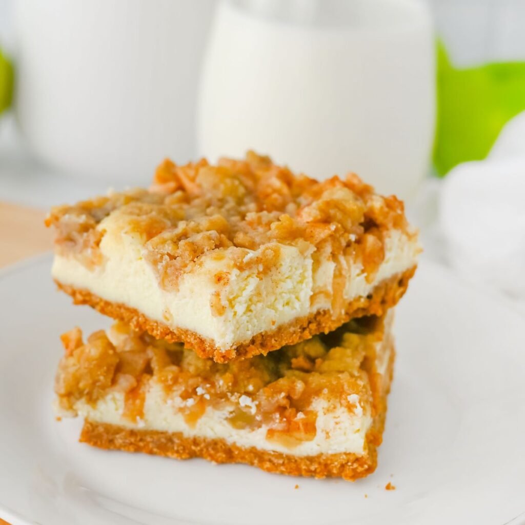 Two crumb-topped cheesecake bars are stacked on a white plate, with a blurred glass of milk and green background visible behind them.