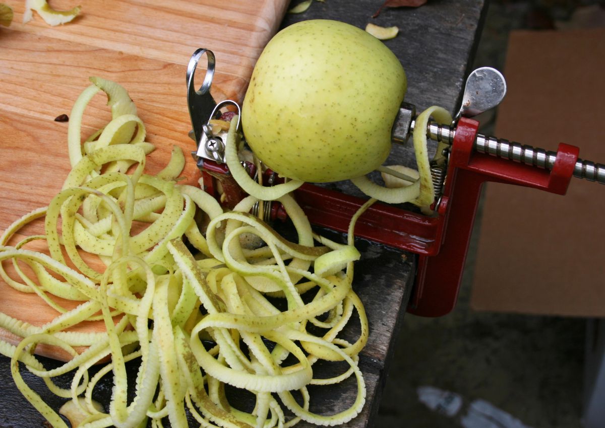 A green apple is being peeled using a mechanical apple peeler, with long strips of apple peel collected on the table.