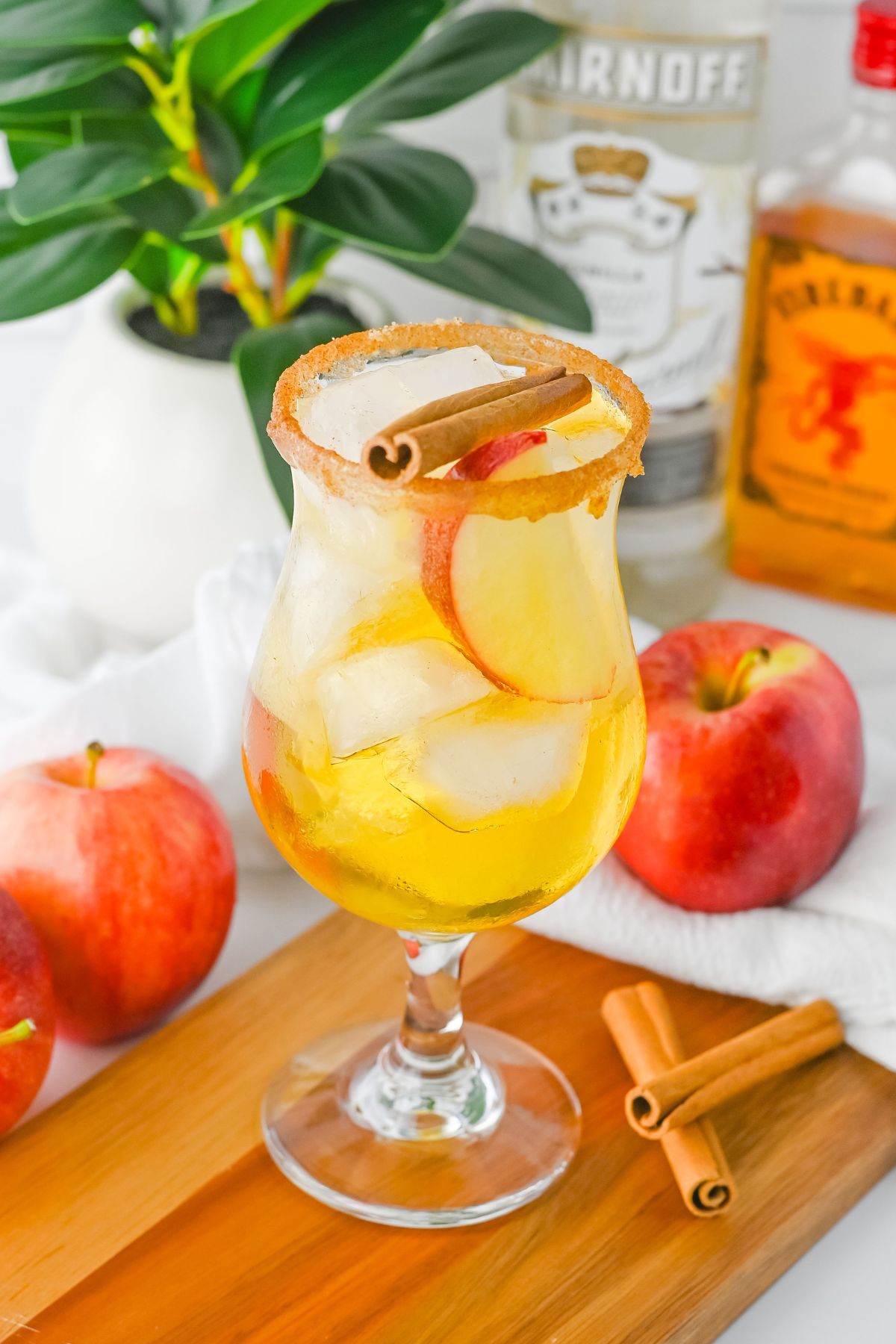 A glass of iced apple cocktail garnished with apple slices and a cinnamon stick, with apples, cinnamon sticks, and liquor bottles in the background.