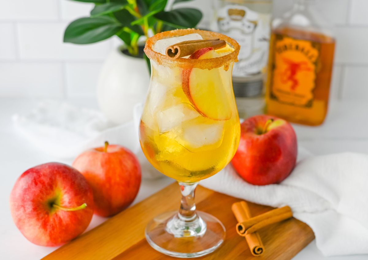 A glass of yellow cocktail with ice, apple slices, and a cinnamon stick, garnished with a cinnamon sugar rim, sits on a wooden board with apples and cinnamon sticks nearby.