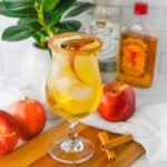 A glass of iced apple cocktail garnished with apple slices and a cinnamon stick, surrounded by apples, cinnamon sticks, and liquor bottles on a white surface.