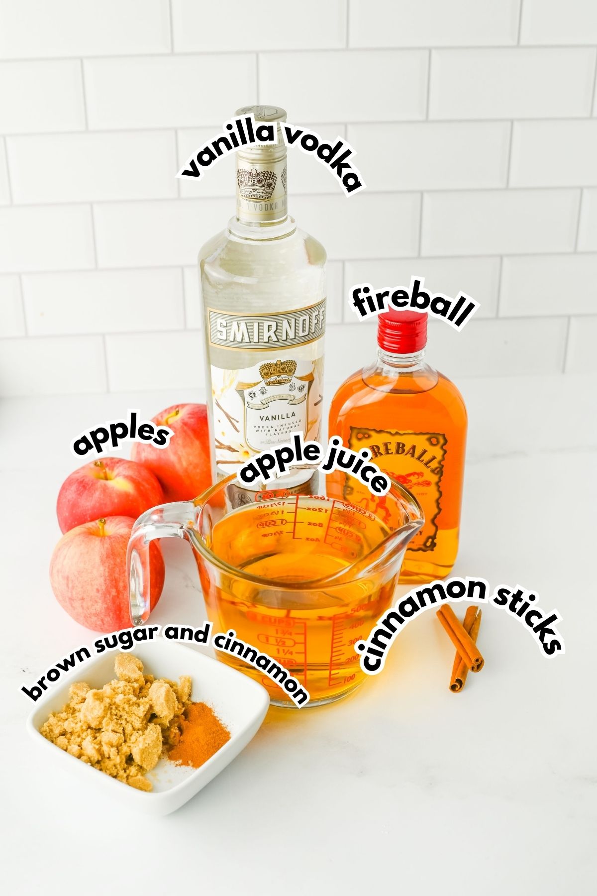 Apples, vanilla vodka, Fireball whiskey, apple juice, brown sugar, cinnamon powder, and cinnamon sticks arranged on a white countertop.