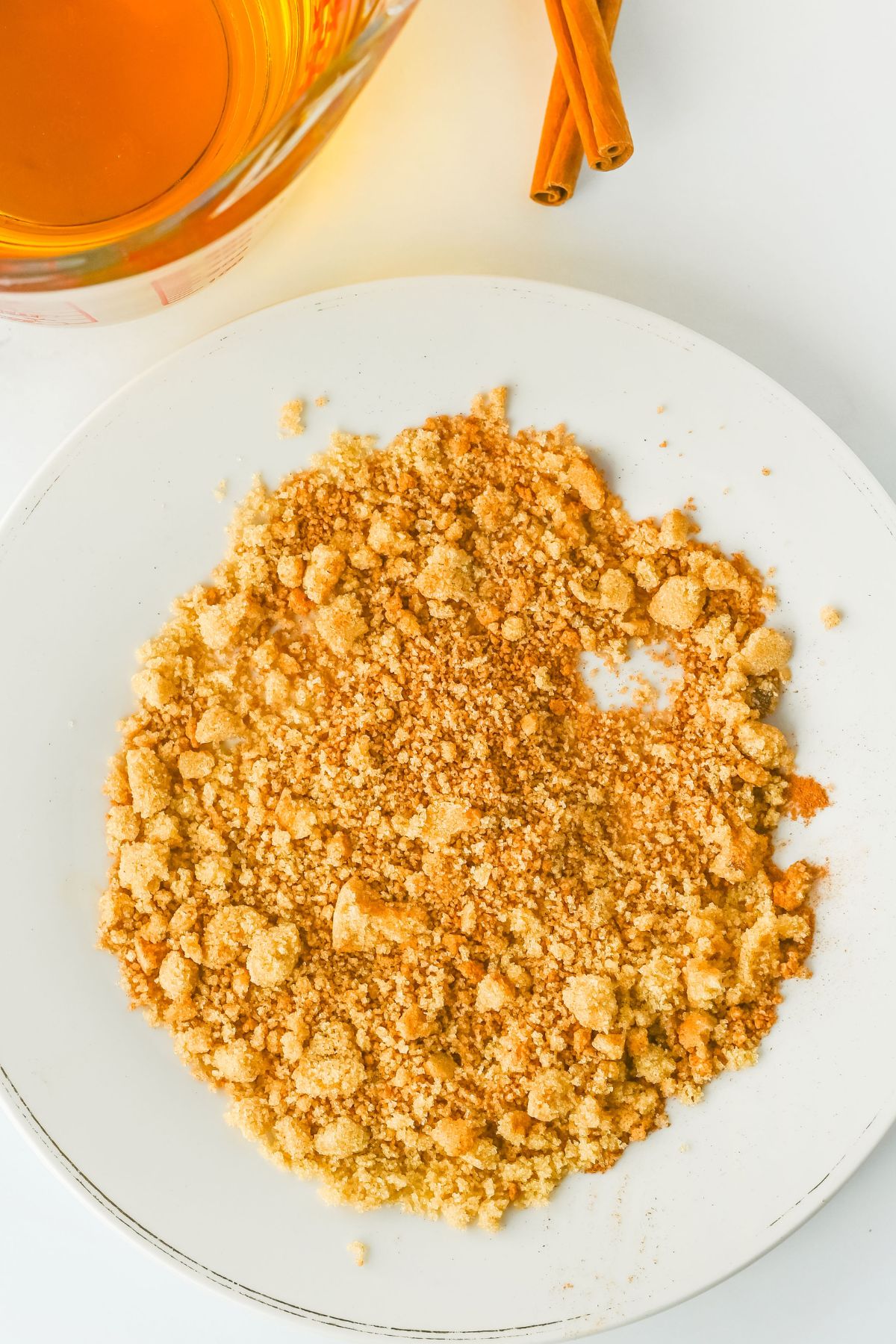 A white plate with a pile of crushed graham crackers sits next to a glass of amber liquid and two cinnamon sticks on a white surface.