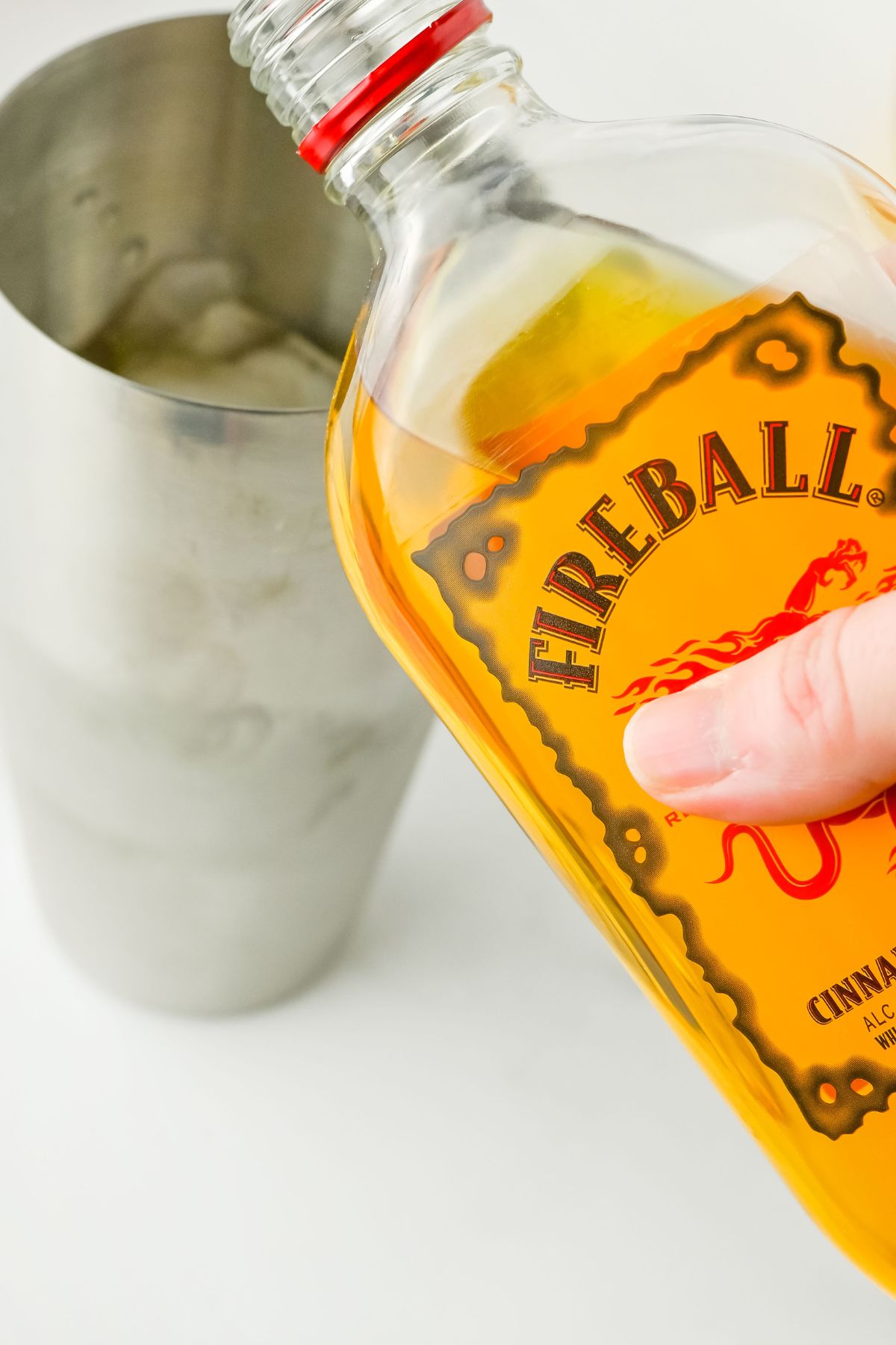 A hand pours Fireball Cinnamon Whisky from a bottle into a metal cocktail shaker filled with ice.