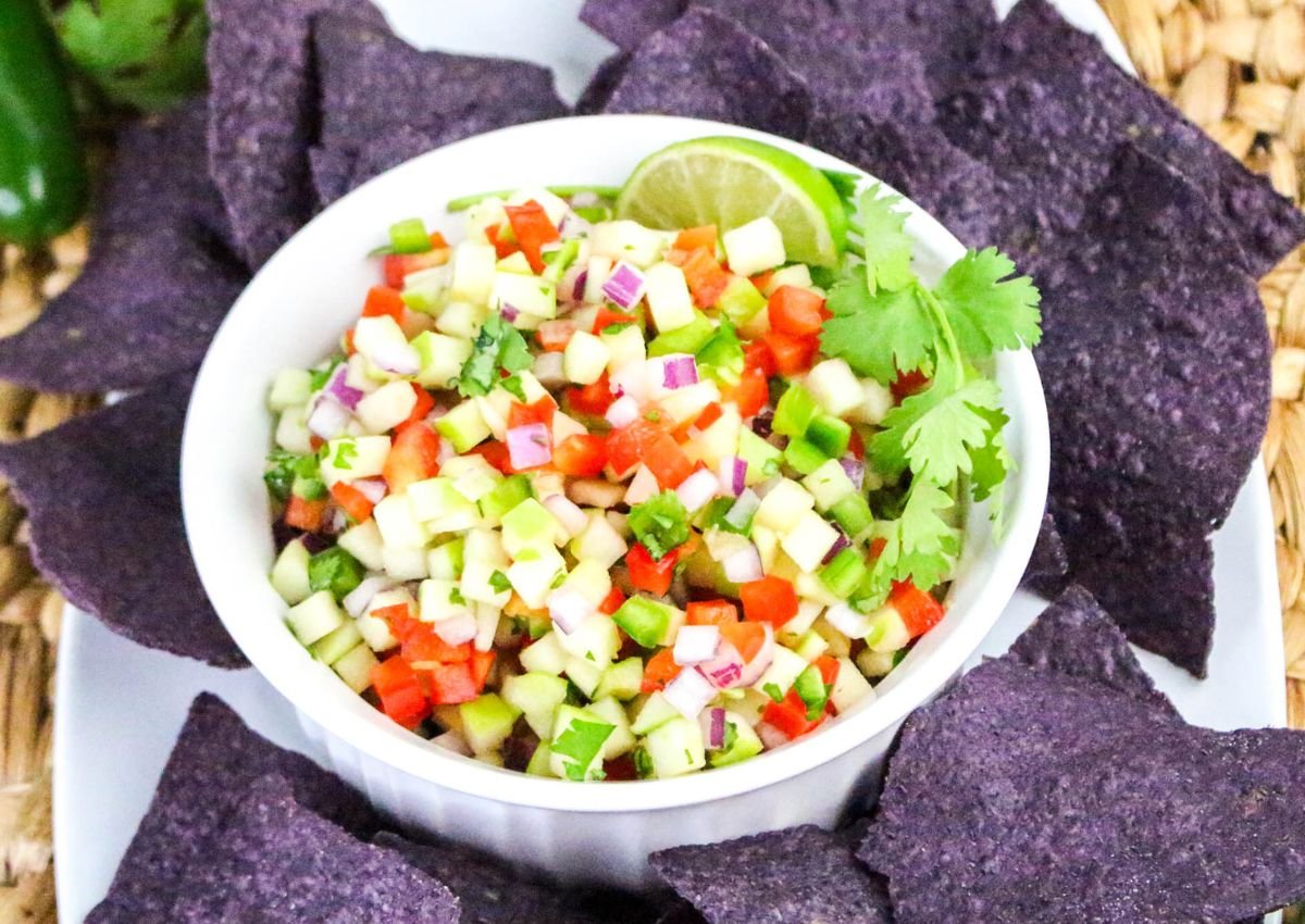 A bowl of diced cucumber salsa garnished with lime and cilantro, surrounded by blue corn tortilla chips on a white plate.