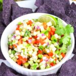 A bowl of fresh cucumber salsa with diced vegetables, garnished with cilantro and a lime slice, surrounded by blue corn tortilla chips.