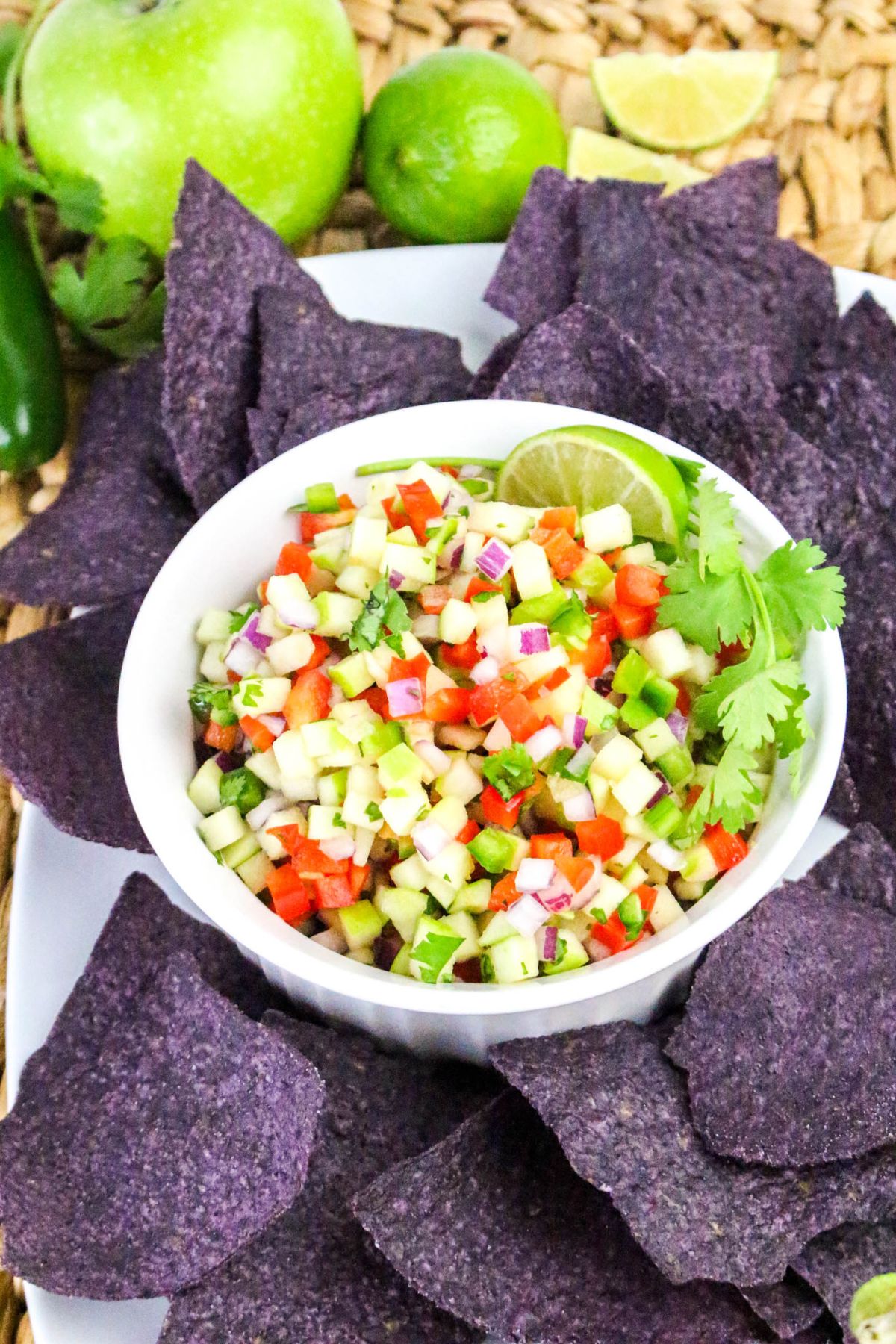 A white bowl filled with diced vegetable salsa, garnished with lime and cilantro, is surrounded by blue corn tortilla chips on a woven surface.