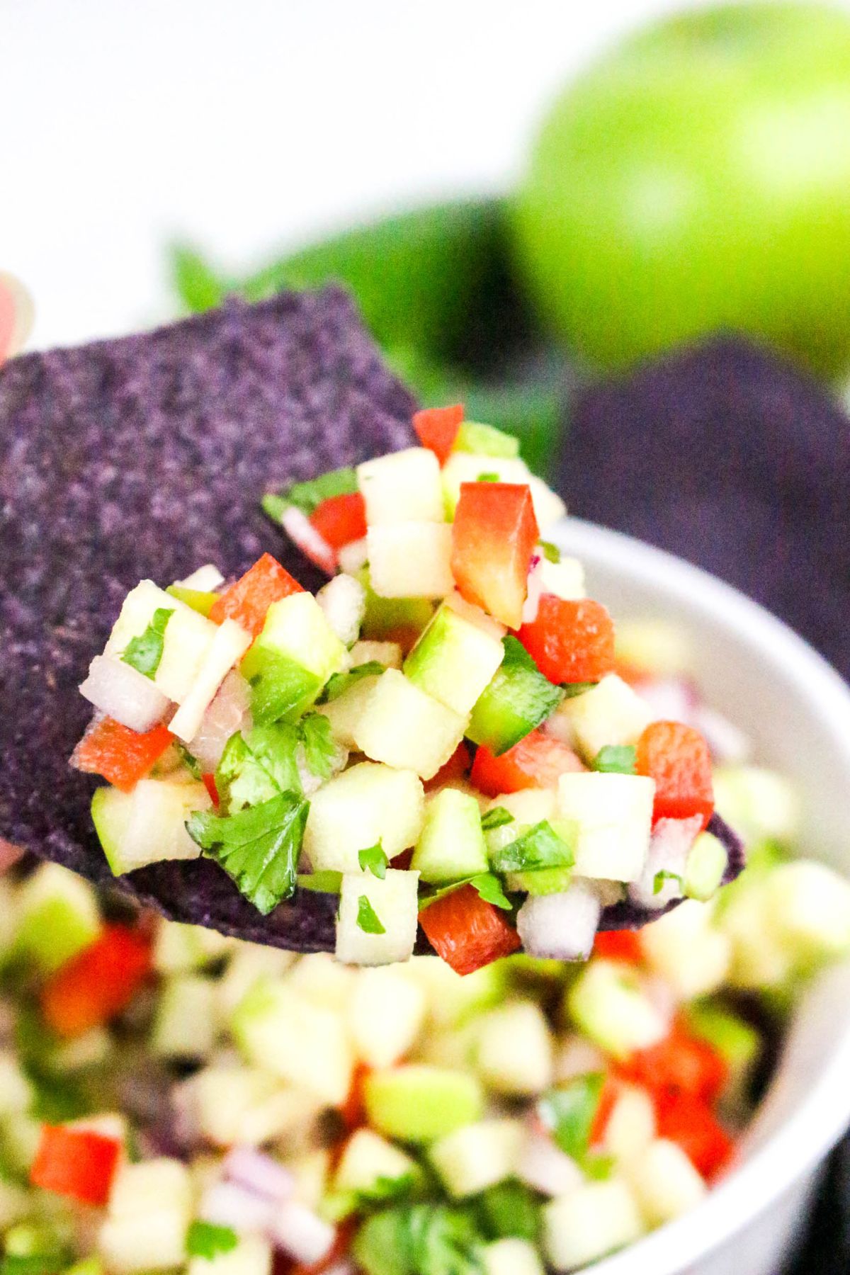 A close-up of a blue corn chip topped with apple salsa containing diced apples, red bell peppers, onions, cilantro, and jalapeño.