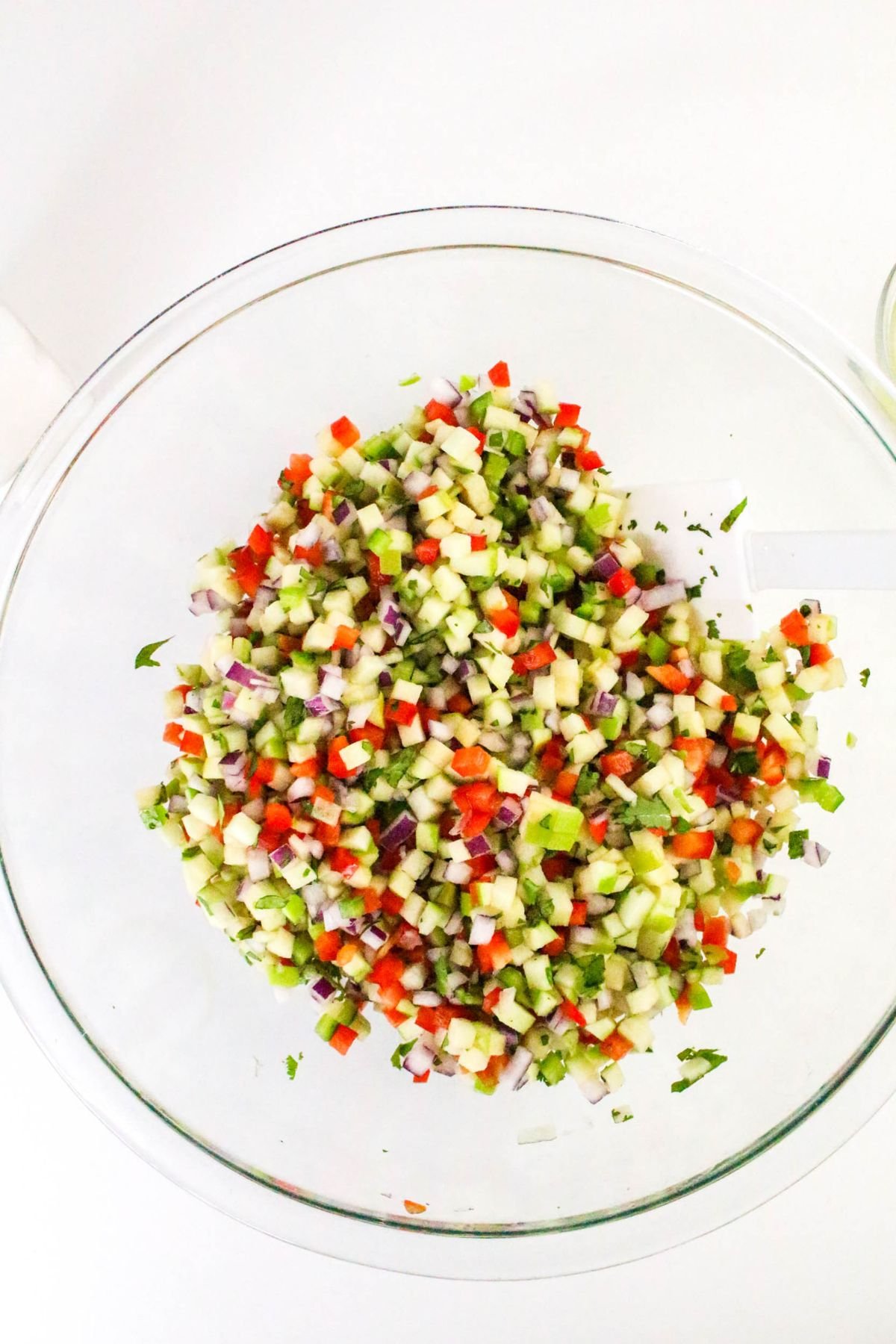 A clear glass bowl filled with finely diced vegetables, including red onion, tomato, cucumber, and green herbs, sits on a white surface with a spatula inside.