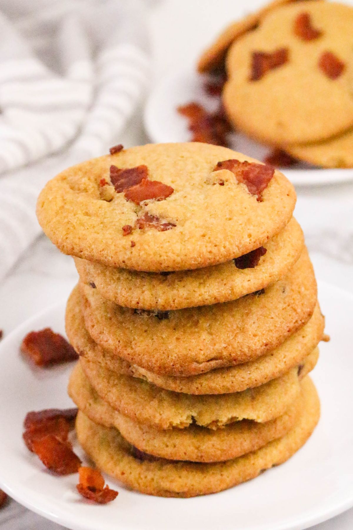 A stack of six cookies with visible bacon pieces sits on a white plate, with more cookies and bacon pieces in the background.