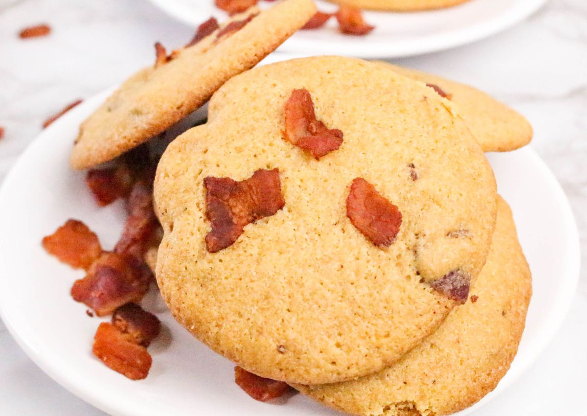 A white plate with cookies topped and mixed with pieces of cooked bacon, with extra bacon pieces scattered around.