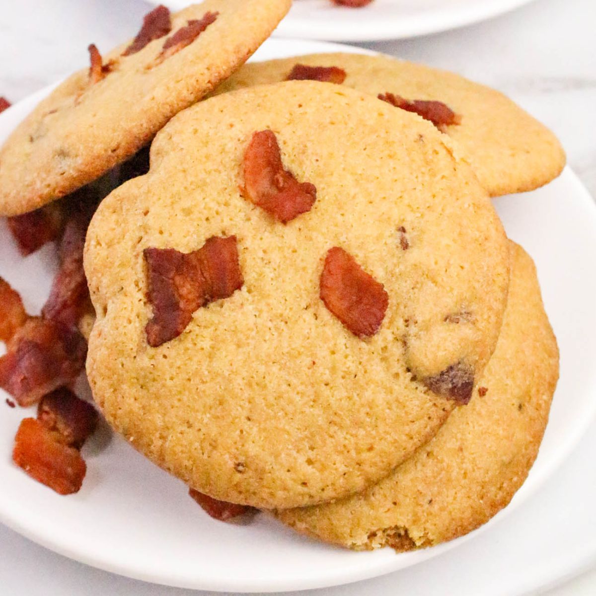 Three cookies topped with pieces of cooked bacon are stacked on a white plate, with additional bacon pieces scattered nearby.