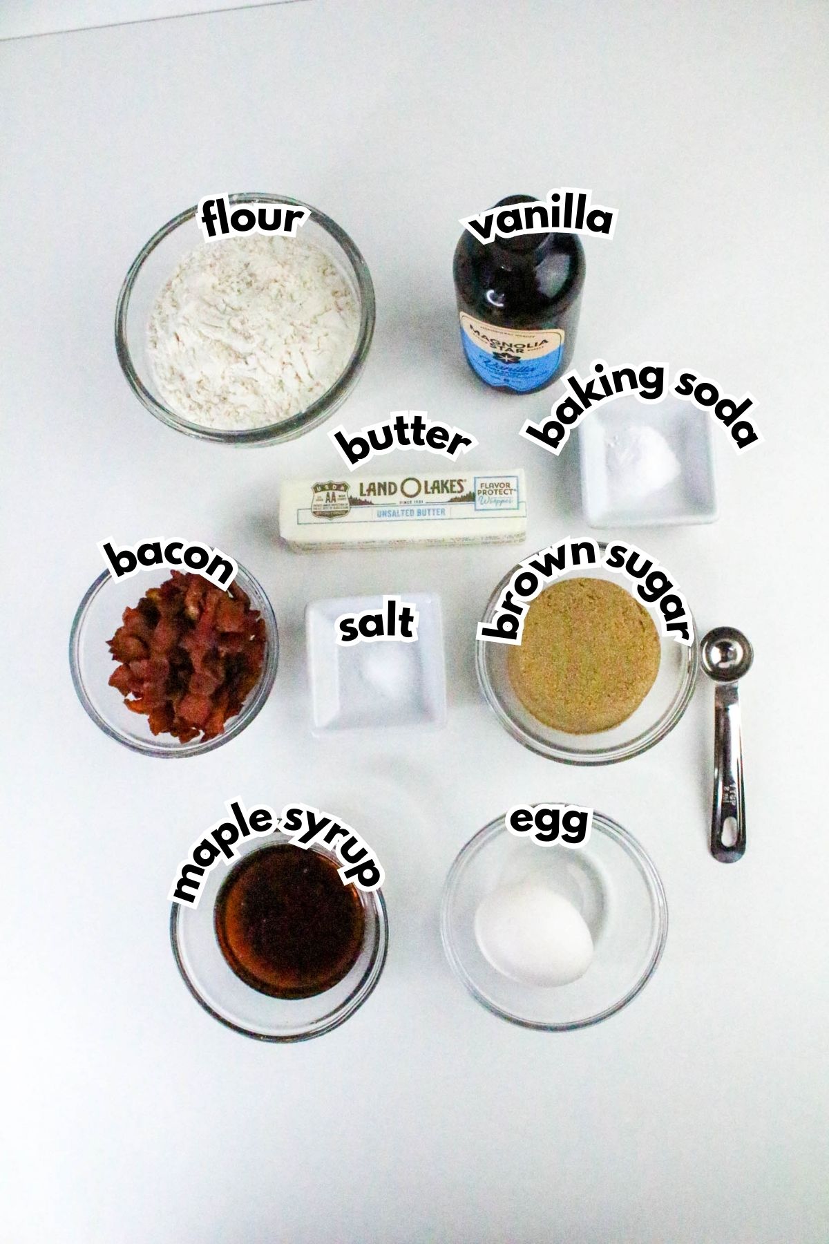 Nine baking ingredients on a white background: flour, vanilla, baking soda, butter, bacon, salt, brown sugar, maple syrup, and an egg, each labeled with text.