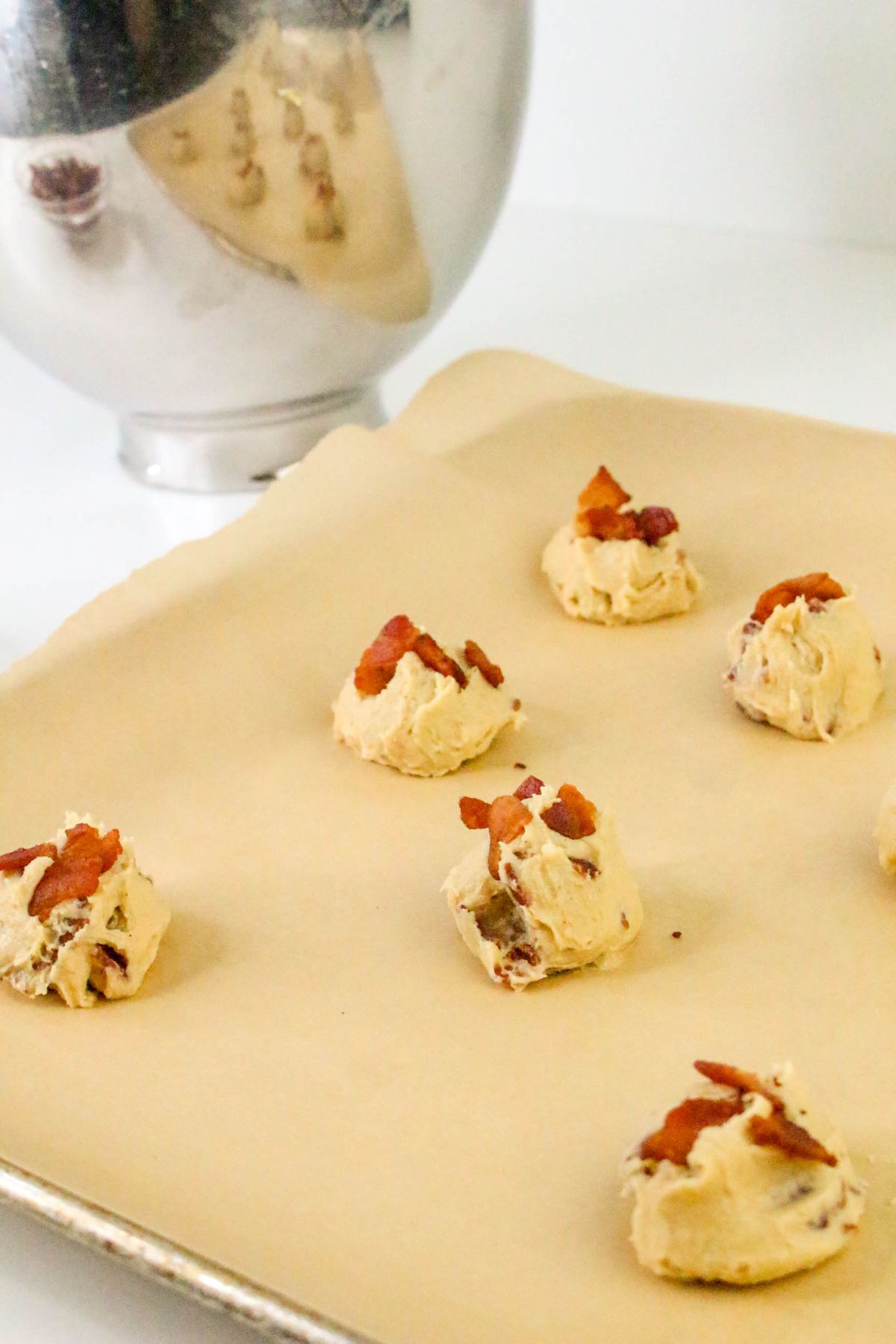 Scoops of cookie dough with bacon pieces on top are spaced on a parchment-lined baking sheet; a metal mixing bowl is in the background.