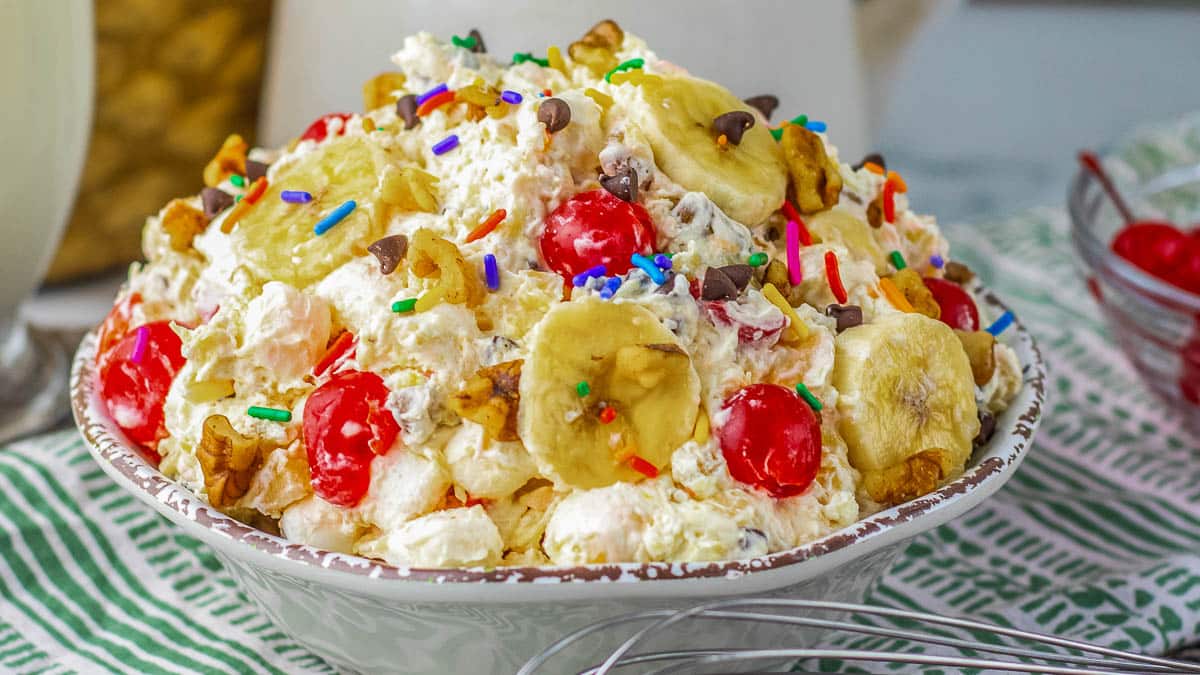 A bowl of ambrosia salad topped with banana slices, cherries, walnuts, sprinkles, and chocolate chips, placed on a striped green and white cloth.