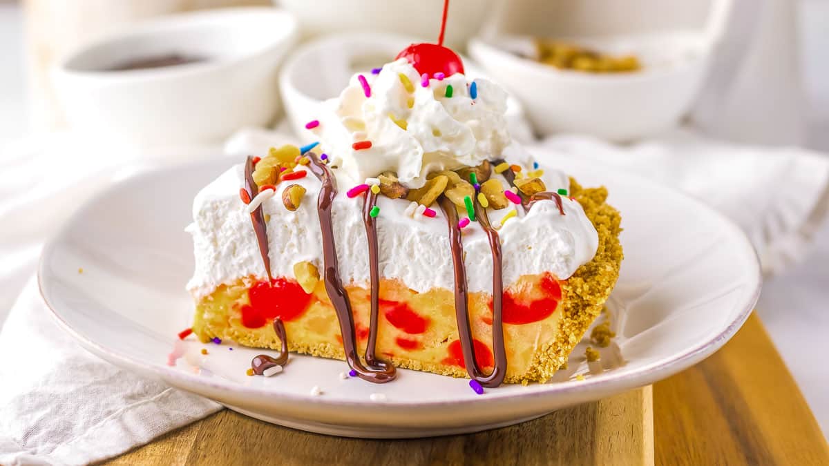 Slice of pie with whipped cream, chocolate drizzle, sprinkles, nuts, and a cherry on top, served on a white plate.