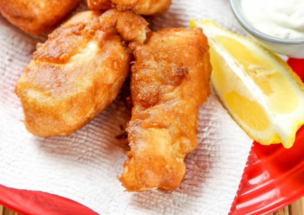 Golden fried fish fillets on a paper towel with a lemon wedge and a small bowl of white dipping sauce on a red plate.