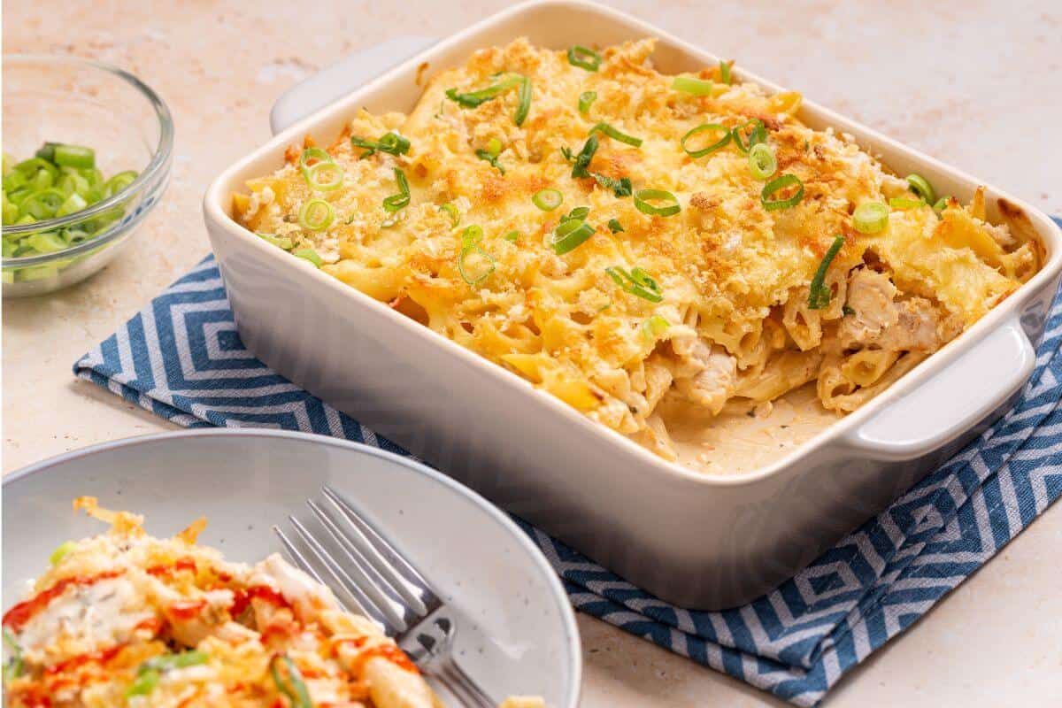 A baked pasta casserole topped with chopped green onions sits in a square dish on a blue patterned cloth, with a serving on a plate beside it.