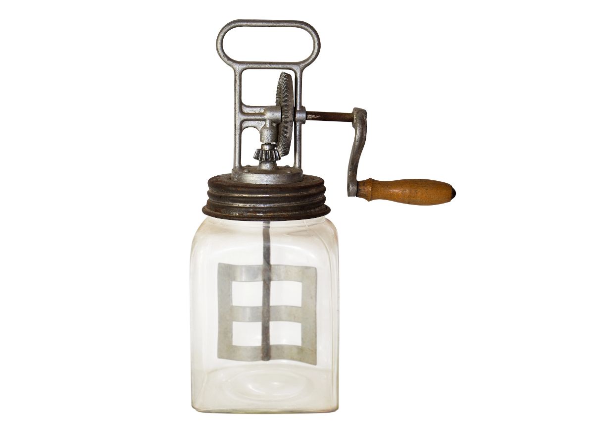 A vintage glass butter churn with a metal gear mechanism and a wooden handle, isolated on a white background.