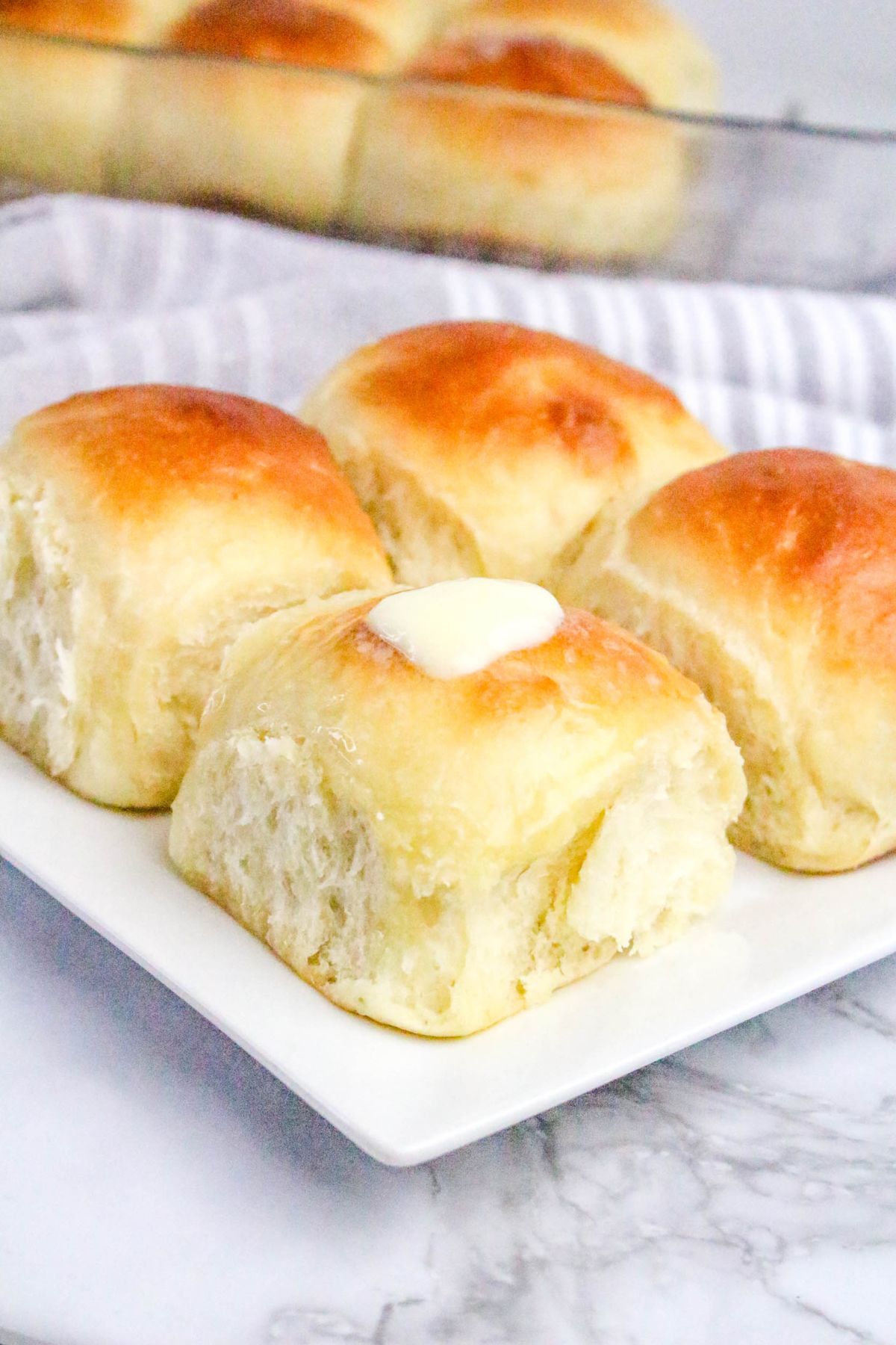 Four golden brown dinner rolls are arranged on a white plate, with a pat of butter melting on top of one roll. More rolls are visible in the background.