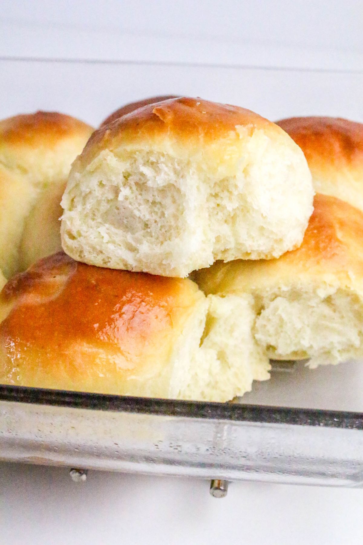 A glass dish containing several golden-brown, fluffy bread rolls, with one roll partially bitten to show its soft texture inside.