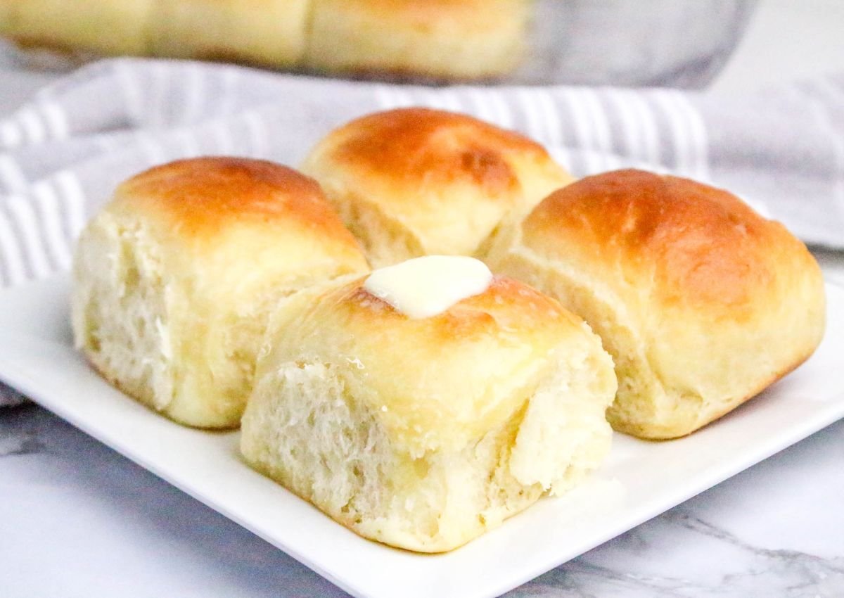 Four golden brown dinner rolls on a white plate, with a pat of butter melting on top of one roll.