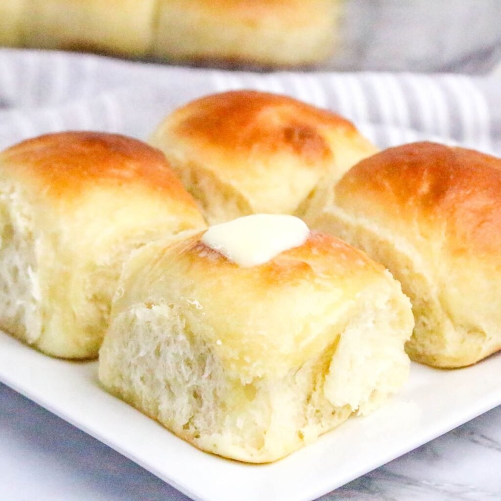 Four golden brown dinner rolls sit on a white plate, with a pat of butter melting on the roll in the foreground.