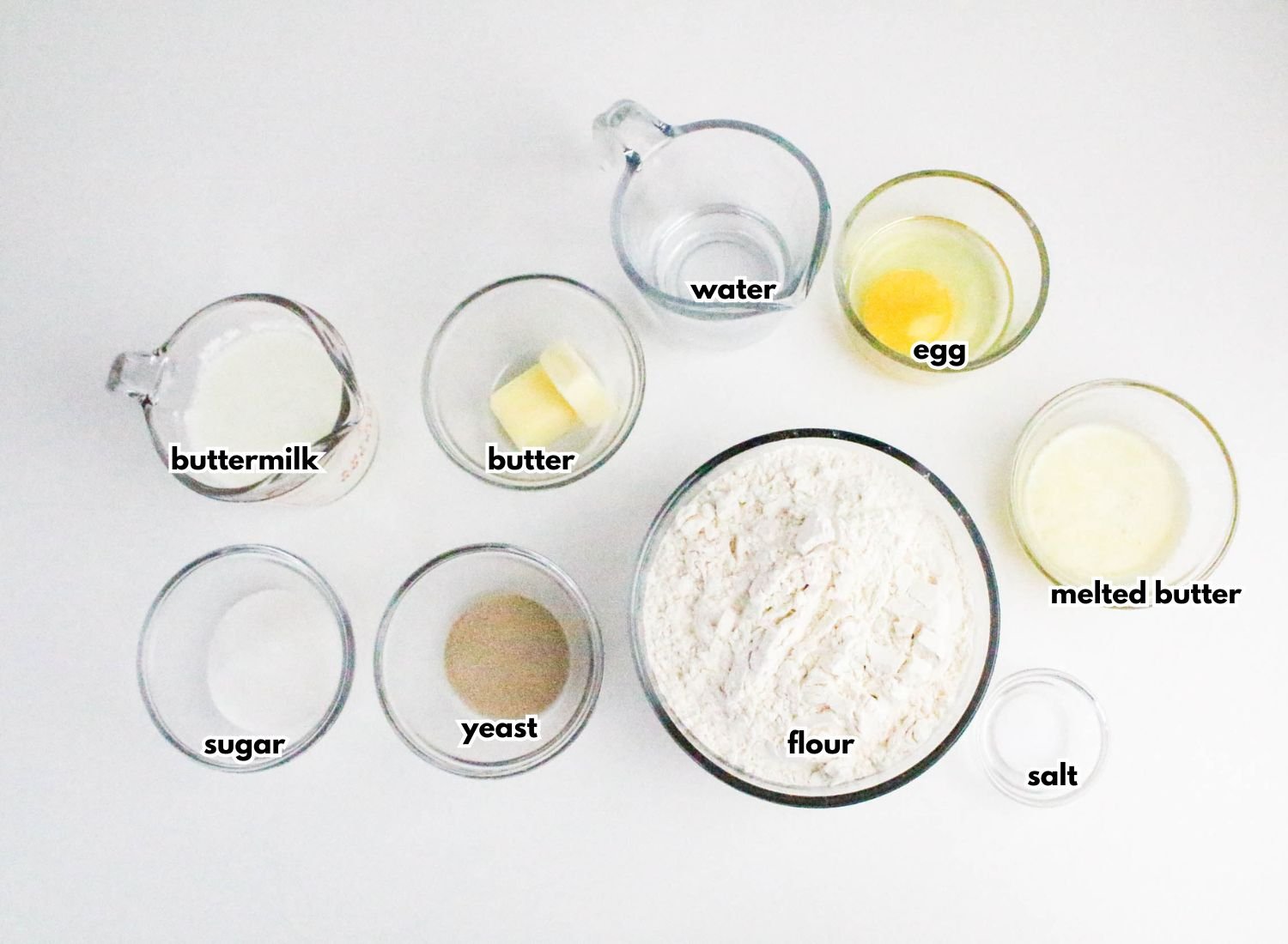 Nine glass bowls containing buttermilk, sugar, yeast, butter, water, egg, flour, melted butter, and salt are arranged on a white surface. Each ingredient is labeled.